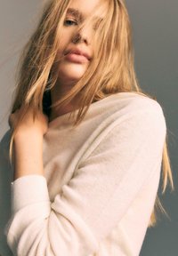 Detailný záber osoby s dlhými blond vlasmi, ktoré čiastočne zakrývajú tvár, oblečenej v svetlom mäkkom svetre, na jednoduchom šedom pozadí.