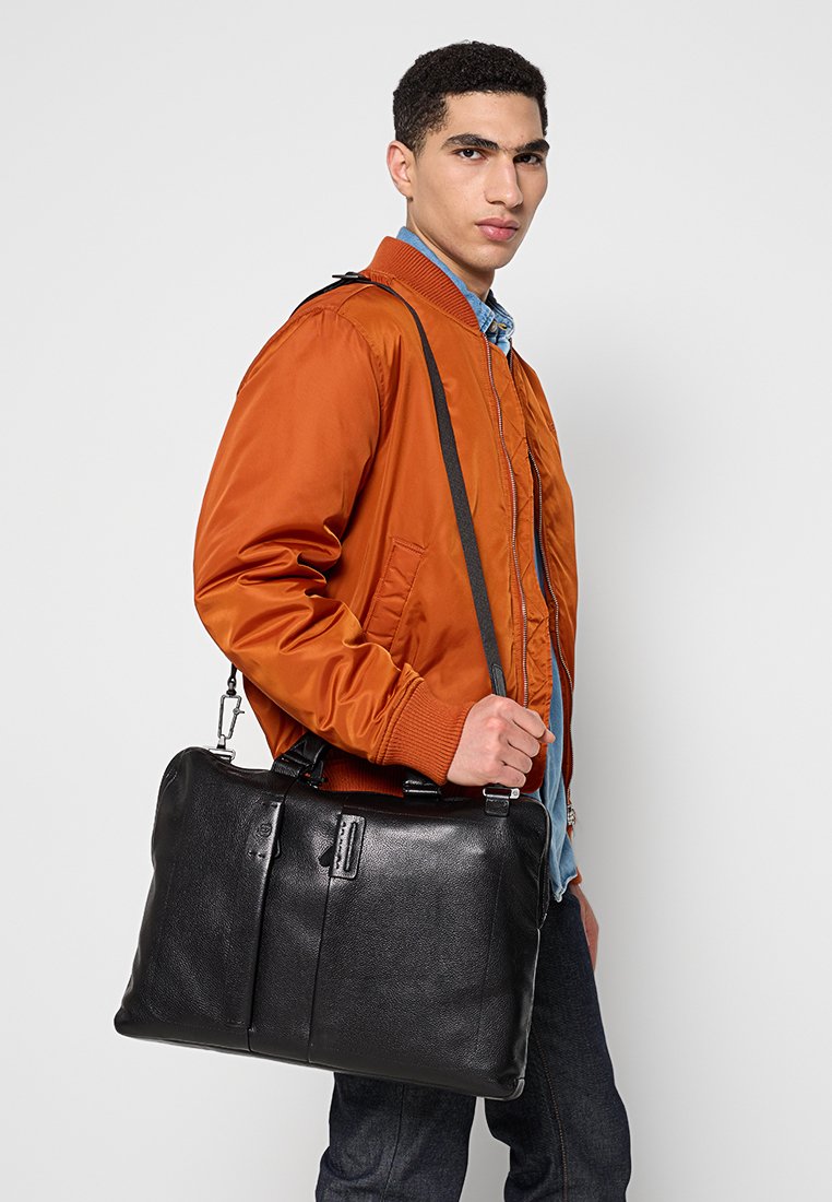 Czarna skórzana torba na laptopa z podwójnymi przednimi kieszeniami na zamek, gładką teksturą, płaskim kształtem i regulowanym paskiem na ramię, trzymana przez osobę w pomarańczowej kurtce.