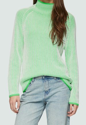 Žena ve svetru s dlouhými rukávy s vertikálními zelenobílými pruhy, zastrčeném do světle modrých džínů, s dlouhými hnědými vlasy přes jedno rameno.