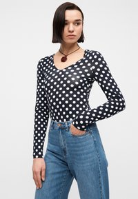 Γυναίκα που φοράει μαύρο μακρυμάνικο τοπ με λευκές πουά, μπλε τζιν ψηλόμεσο και κολιέ με μενταγιόν κόκκινο τριαντάφυλλο, στέκεται με το ένα χέρι στην τσέπη.