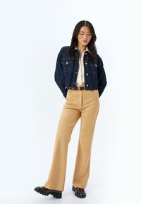 Chaqueta de mezclilla corta con cuello puntiagudo, botones dorados y dos bolsillos en el pecho. Pantalones de pierna ancha en color beige con costuras verticales y cinturón. Zapatos de plataforma negros.