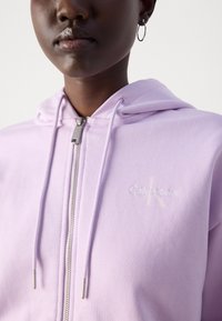 Šviesiai violetinė užtrauktuku užsegama megztinė, pagaminta iš minkštos medžiagos su raišteliais ir sidabro spalvos detalėmis; turi išsiuvinėtą "Calvin Klein" logotipą derančia spalva.
