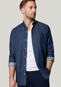 Jeansowa koszula zapinana na guziki w ciemnoniebieskim kolorze, z podwiniętymi rękawami, jasnoniebieskimi mankietami i białymi guzikami. Nosi się ją na białym podkoszulku.