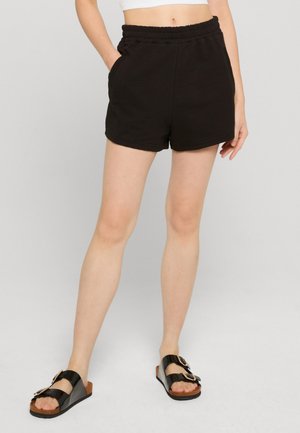Femme portant un short texturé noir et des sandales compensées noires, debout avec une main dans la poche sur un fond clair.