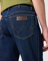 Los pantalones vaqueros de mezclilla azul oscuro presentan un parche de cuero de Wrangler y una costura distintiva en los bolsillos traseros en un patrón curvado. Tela texturizada visible.