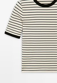 Kurzärmliges cremefarbenes Hemd mit dünnen schwarzen horizontalen Streifen und schwarzem Besatz am Halsausschnitt und an den Bündchen.