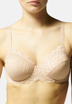 Kobieta w beżowym koronkowym staniku z kwiatowymi wzorami i regulowanymi ramiączkami, widoczna od ramion do górnej części tułowia.