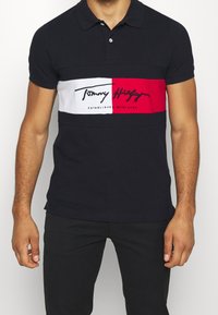Camisa polo azul marino con una franja horizontal blanca y roja que presenta texto negro. Mangas cortas, cuello clásico y un ajuste relajado.