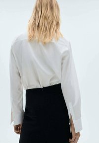 White long-sleeve button-up shirt with a smooth texture, features a back vent, worn over a high-waisted black skirt.