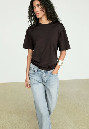 Kobieta o kręconych włosach, ubrana w czarny t-shirt z krótkim rękawem, jasnoniebieskie dżinsy i srebrną bransoletkę, stoi z jedną ręką w kieszeni.
