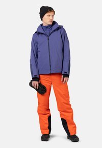 Fioletowa wodoodporna kurtka z wysokim kołnierzem i czarnymi akcentami, w połączeniu z jasnopomarańczowymi spodniami z czarnymi detalami oraz czarnymi rękawicami.
