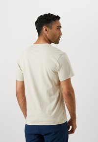 T-shirt en coton beige à manches courtes, col rond et texture lisse, portée par une personne faisant légèrement face sur le côté.