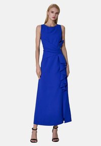 Femme portant une robe sans manches de couleur bleu royal, longueur cheville, avec des volants sur un côté, associée à des talons à bride de cheville noirs.