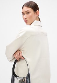 Svetlo béžová, prehnaná bunda z umelého kože so hladkou textúrou. Model drží lesklú striebornú tašku s čiernym popruhom.