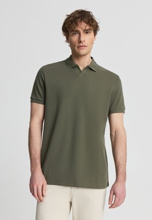 Uomo con capelli ricci che indossa una polo verde oliva a maniche corte e pantaloni beige chiaro, in piedi davanti a uno sfondo neutro.