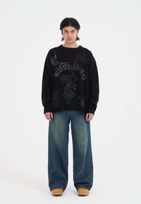 Maglione a coste nero con grafiche. Abbinato a jeans larghi blu sbiaditi e zoccoli color cammello. Outfit semplice e informale con textures distinte.