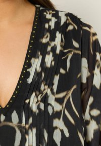 Zwarte blouse met een bloemenpatroon in beige, voorzien van plooien en een V-hals. Gouden kralen accentueren de halslijn voor detail.