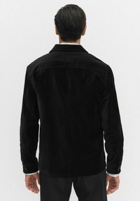 Zwarte corduroy jas met een ontspannen pasvorm, zichtbare stiksels en manchetten met knopen. Soepele textuur en klassieke kraagdetail.