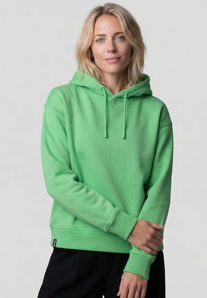 Blonde Frau mit blauen Augen, die einen leuchtend grünen Hoodie und schwarze Hosen trägt, steht mit leicht verschränkten Armen vor einem schlichten Hintergrund.