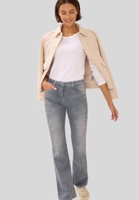 Beige wollen blend jas met grote knopen, gecombineerd met een wit overhemd met lange mouwen en lichtblauwe flared jeans. Lichte sneakers maken de outfit compleet.