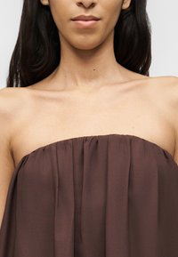 Γυναίκα με μακριά σκούρα μαλλιά, που φοράει ένα αμάνικο, πλισέ καφέ τοπ, απεικονίζεται από τους ώμους έως λίγο κάτω από το πηγούνι.