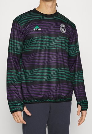 Czarna sportowa koszulka z długim rękawem z zielonymi i fioletowymi ukośnymi paskami, logo Adidas i herb Realu Madryt, noszona przez osobę.