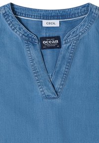 Chemise en jean bleu clair avec col en V et deux étiquettes cousues, l'une indiquant "CECIL" et l'autre avec les phrases "océan," "eau salée & soleil," et "brise fraîche."