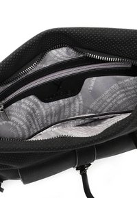 Innenraum einer schwarzen Tasche mit perforierter Textur, silbernem Reißverschluss und grauem, gemustertem Futter mit Text, sichtbar ist ein Markenlogo auf der Tasche.