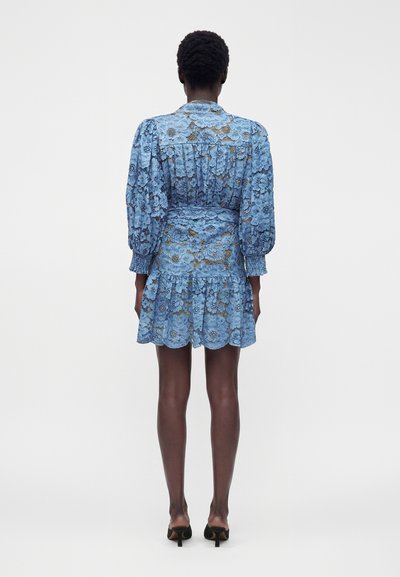 Femme portant une robe en dentelle bleue longueur genou, à manches longues, avec des motifs floraux et des manches bouffantes, vue de dos sur un fond uni.