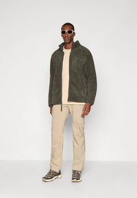 Zelena jopa iz flisa z zadrgo spredaj, bež majica, bež hlače s stranskimi žepi in temna obutev. Dodatki vključujejo svetleče sončna očala.