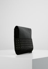 Černá kožená taška s klopovým uzavíráním, která má texturovaný černý látkový pruh zdobený malými stříbrnými cvočky. Kompaktní obdélníkový tvar.