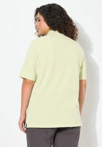 Bledě žlutá košile s krátkým rukávem, má límec, boční rozparky a hladkou texturu. Nosená s šedými kalhotami, zobrazující pohled zezadu.