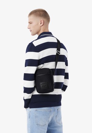 Bolso cruzado negro con parche delantero texturizado, llevado sobre un suéter de rayas azul marino y blanco, combinado con jeans de mezclilla azul claro.