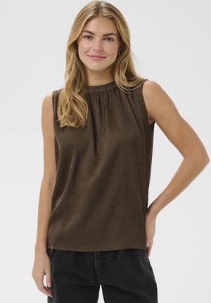 Femme aux cheveux blonds portant un haut sans manches marron foncé texturé et un pantalon noir, souriante, une main sur la hanche.