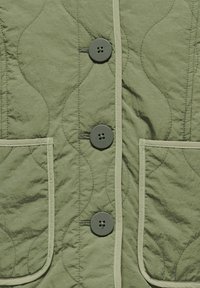 Grön quiltad jacka med knappstängning, som har fyra knappar, två fickor och kontrastrande ljus sömnad längs kanterna.