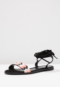 Black suede sandal with pink and white beaded strap. Features lace-up design around ankle and flat sole.