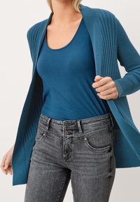 Teal ribbad cardigan över en teal linne, tillsammans med grå jeans med tvåknappsslutning och sydda fickdetaljer.