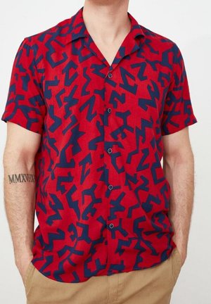 Uomo con camicia rossa a maniche corte con motivo geometrico astratto blu navy e pantaloni color sabbia, mani in tasca.