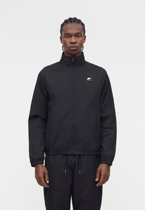 Mladý muž s vrkočmi, oblečený v čiernu bundu Nike Air so zipsom a čierne nohavice, stojaci pred jednoduchým bielym pozadím.