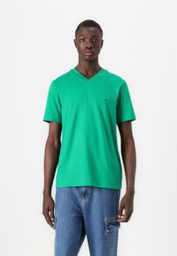 T-shirt vert en coton avec col en V et manches courtes, comportant un petit logo sur la poitrine. Coupe droite et texture douce. Porte avec un pantalon en denim bleu.