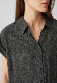 Kurzärmliges Button-up-Hemd aus dunkelgrünem Stoff mit einem strukturierten, geometrischen Muster. Verfügt über einen Kragen und schwarze Knöpfe.