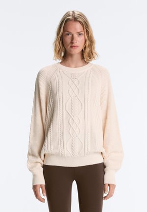 Femeie cu păr blond până la umeri, purtând un pulover tricotat crem și pantaloni maro închis, privindu-se înainte pe un fundal simplu.