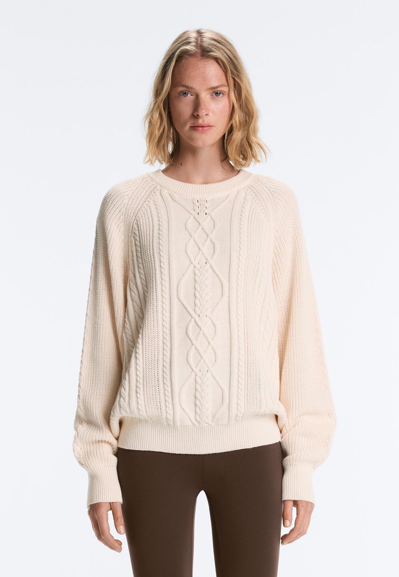 Femme aux cheveux blonds mi-longs portant un pull torsadé couleur crème et un pantalon marron foncé, face à un fond uni.
