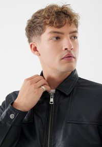 Giubbotto in pelle nera con colletto eretto, chiusura con zip e dettagli con bottoni a pressione. Tessuto liscio, con una tasca sul petto e cuciture alla moda.