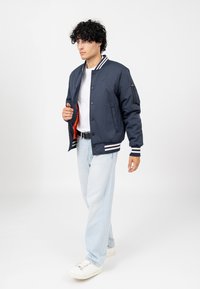 Marine bomberjack met witte gestreepte ribboordmanchetten en kraag, oranje voering, knopenlijst aan de voorkant en zijzak met rits. Gemaakt met lichte denim jeans.