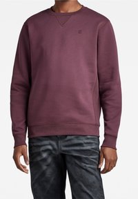 Sudadera marrón con cuello redondo y puños acanalados, que presenta un sutil detalle de logo. Textura suave y corte relajado, combinada con jeans oscuros.