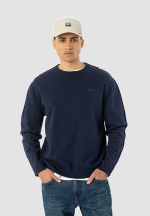 Navyblaues Rundhals-Sweatshirt mit gerippten Bündchen und Saum, ausgestattet mit einem kleinen schwarzen Logo auf der Brust. Kombiniert mit hellblauen Jeans.