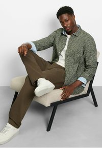 Grünes gemustertes Hemd, hellblaue Denim-Manschetten, weißer Pullover, braune Hose und weiße Schuhe, sitzend auf einem hellbeigen Stuhl mit schwarzen Beinen.