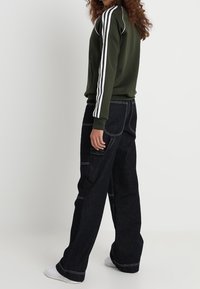 Olivgrön träningsjacka med vit tre-randsdesign, paired med hög midja, vida mörk denimjeans. Vita sneakers fulländar looken.