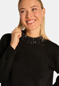 Czarny sweter dzianinowy z ozdobnym kołnierzykiem wyszywanym cekinami. Dekolt okrągły, z subtelnymi akcentami. Materiał wydaje się miękki i teksturowany.